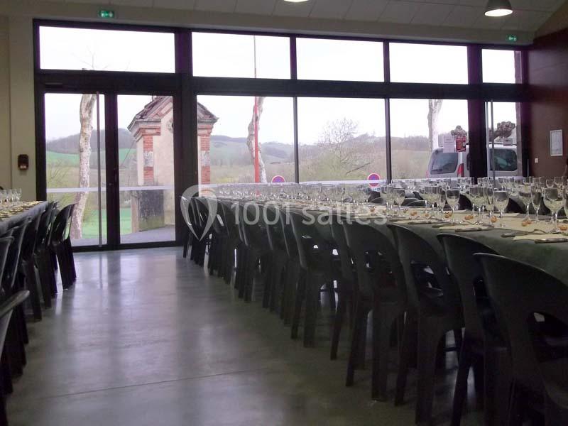 Salle de réception avec de longues tables dressées, chaises noires et vue sur l'extérieur à travers de grandes baies vitrées.
