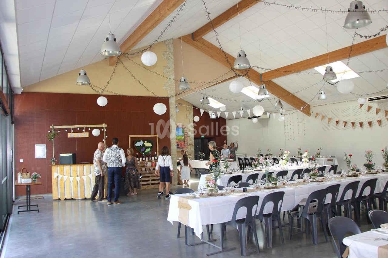 Salle décorée pour un événement, avec tables dressées, guirlandes lumineuses et invités discutant près d'un buffet.