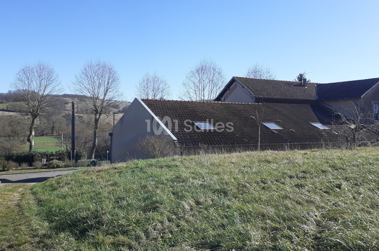 Maison avec toit en tuiles entourée de champs et d'arbres, sous un ciel bleu dégagé.