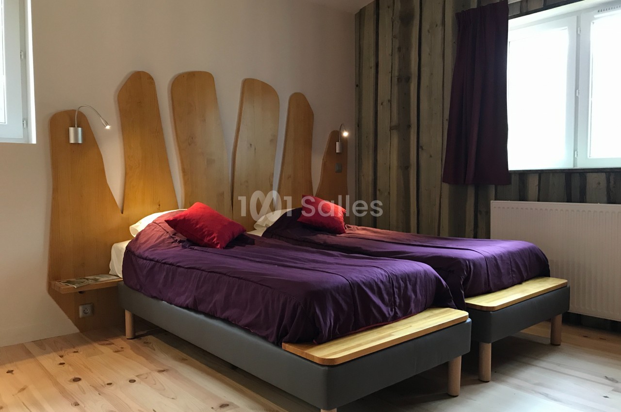 Deux lits simples avec draps violets, têtes de lit en bois sculpté, dans une chambre lumineuse avec parquet et mur en bois.