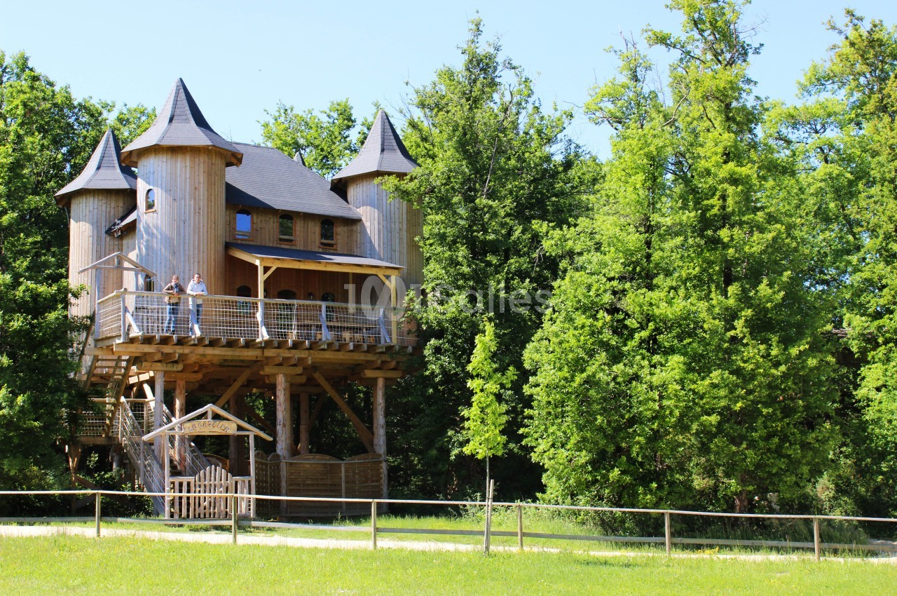 Cabane en bois sur pilotis avec des tours, entourée d'arbres et située dans un espace naturel verdoyant.
