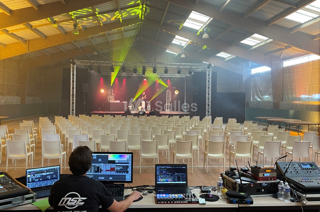 Technicien contrôlant le son et les lumières dans une salle avec scène éclairée et rangées de chaises vides.