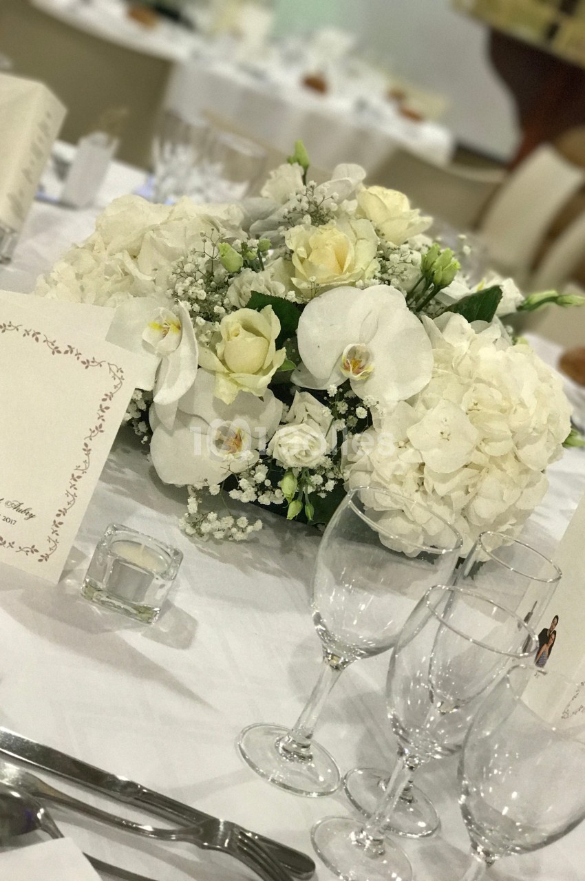 Centre de table floral blanc avec roses, orchidées et hortensias, entouré de verres et menus sur une nappe blanche.