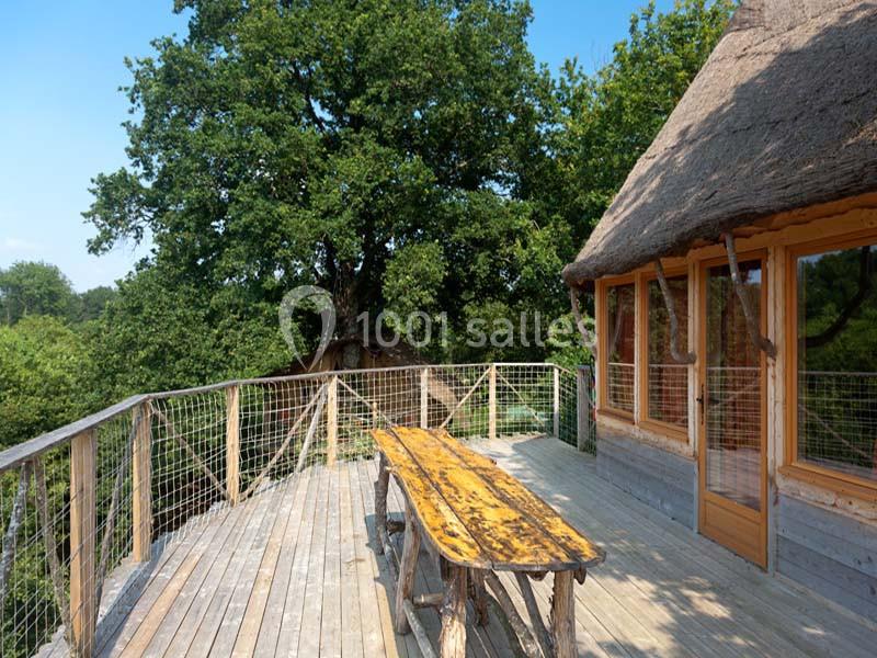 Terrasse en bois avec table et bancs, entourée de verdure, attenante à une cabane au toit de chaume.