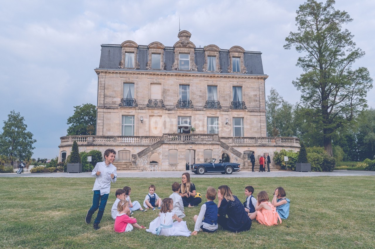 Un groupe d'enfants assis sur une pelouse devant un grand bâtiment ancien, avec des adultes à proximité.