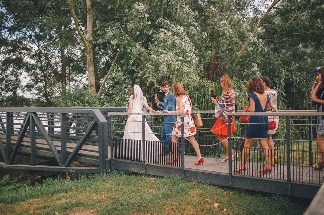 Un couple de mariés traverse un pont en bois, suivi de plusieurs invités vêtus de tenues colorées.