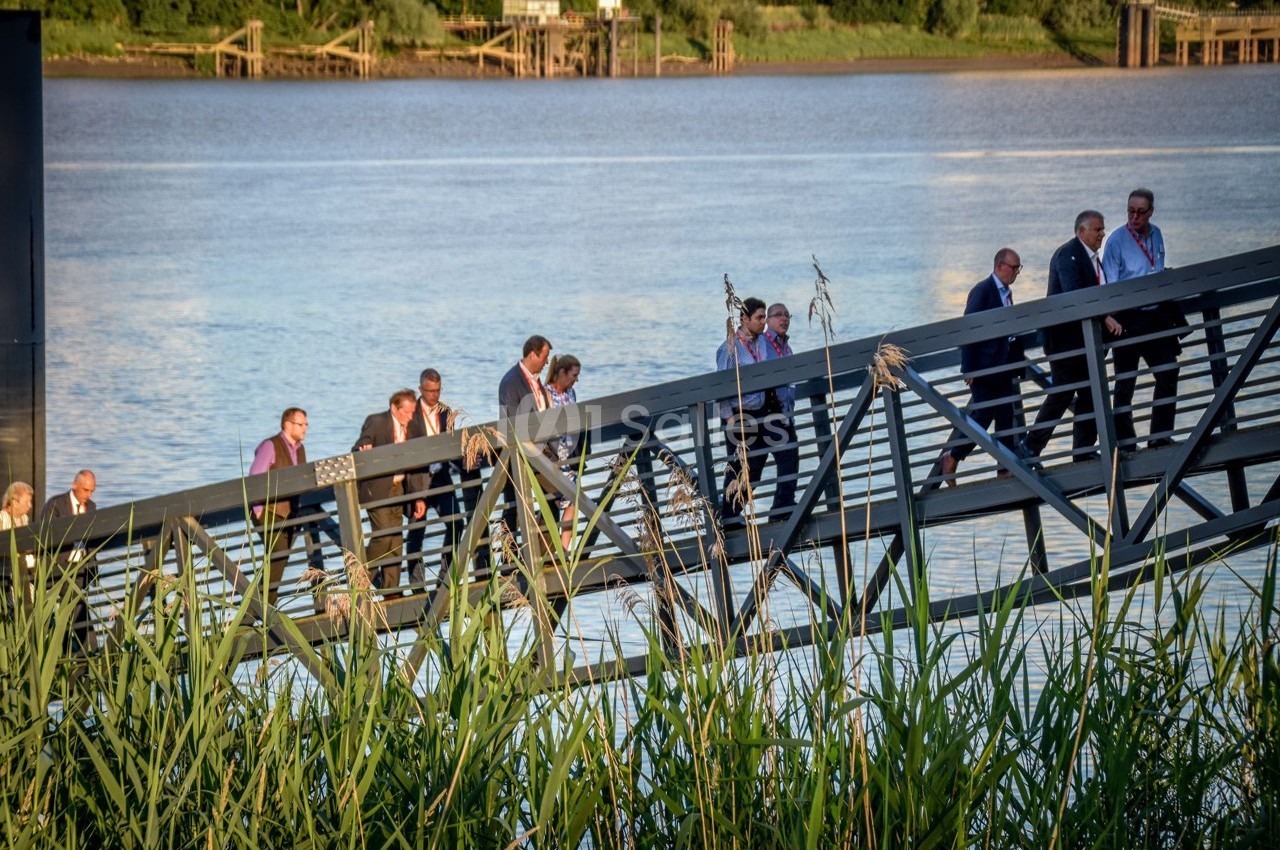 Un groupe de personnes en tenue formelle descend une passerelle métallique au bord d'une rivière.