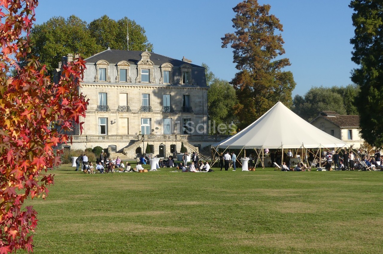 Manoir en pierre entouré d'arbres, avec une tente blanche et des personnes rassemblées sur une pelouse.