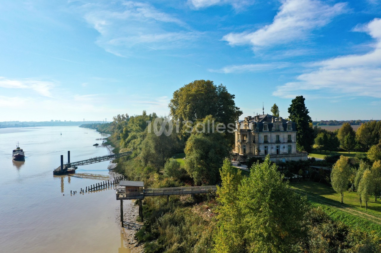 Vue aérienne d'un manoir bordant une rivière, entouré d'arbres et d'un quai avec un bateau à proximité.