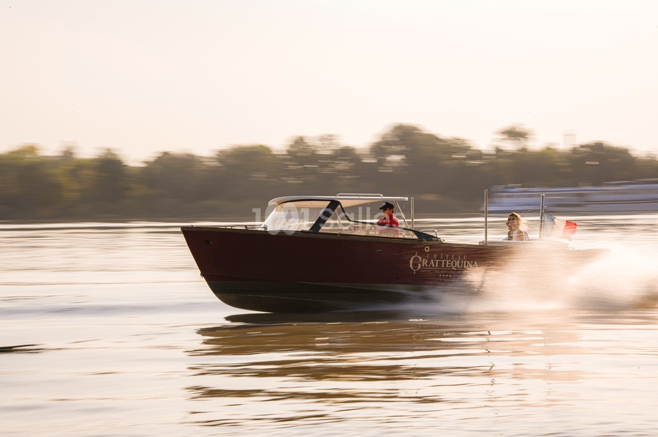 Un bateau à moteur rouge navigue rapidement sur une rivière, avec deux personnes à bord et des arbres en arrière-plan.