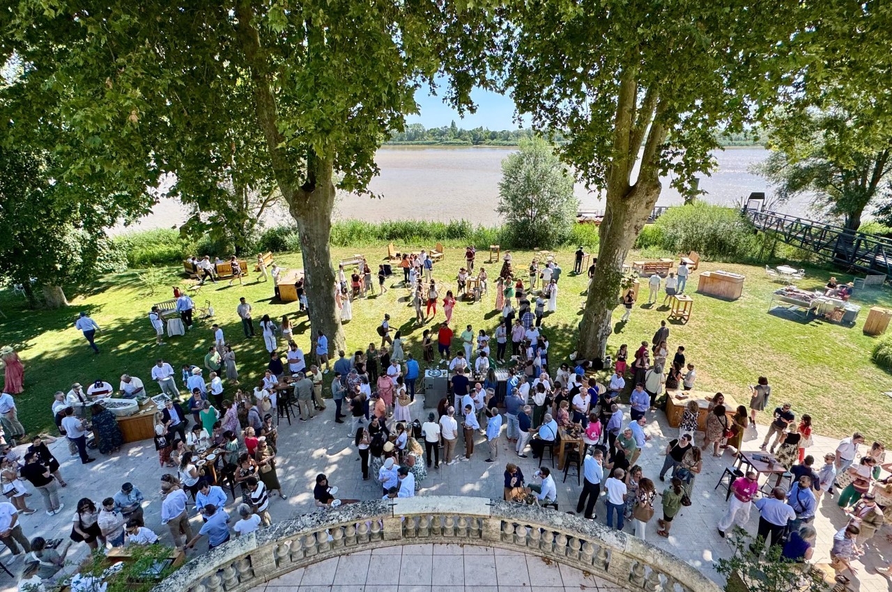 Groupe de personnes rassemblées dans un jardin au bord d'une rivière, entouré d'arbres et de mobilier en plein air.