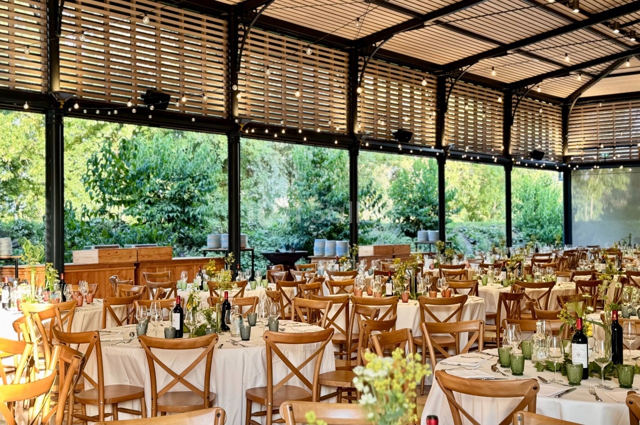 Salle de réception lumineuse avec tables dressées, chaises en bois et vue sur un jardin verdoyant à travers de grandes baies…