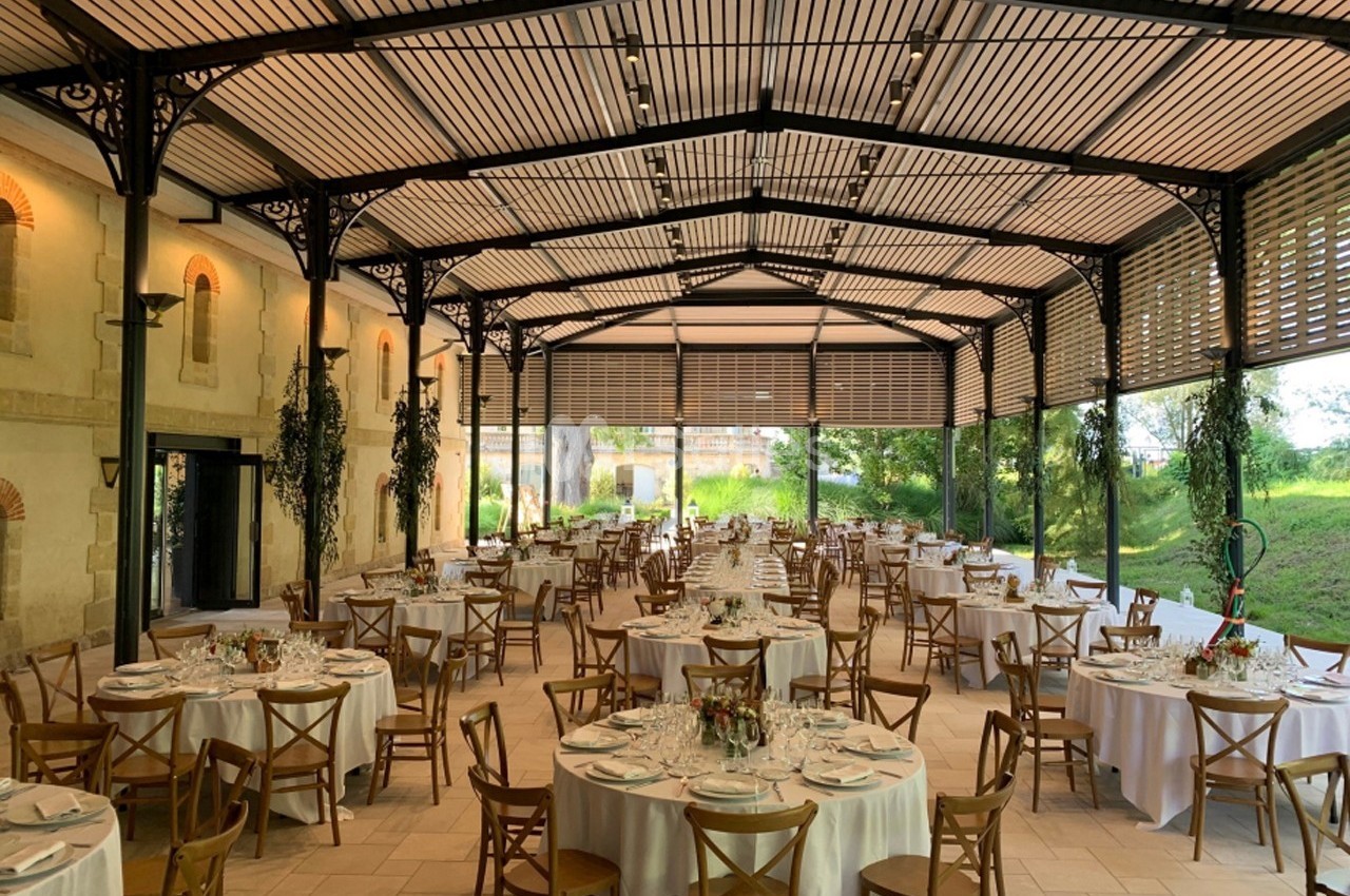 Salle de réception lumineuse avec tables rondes dressées, chaises en bois et grandes baies vitrées donnant sur un jardin.