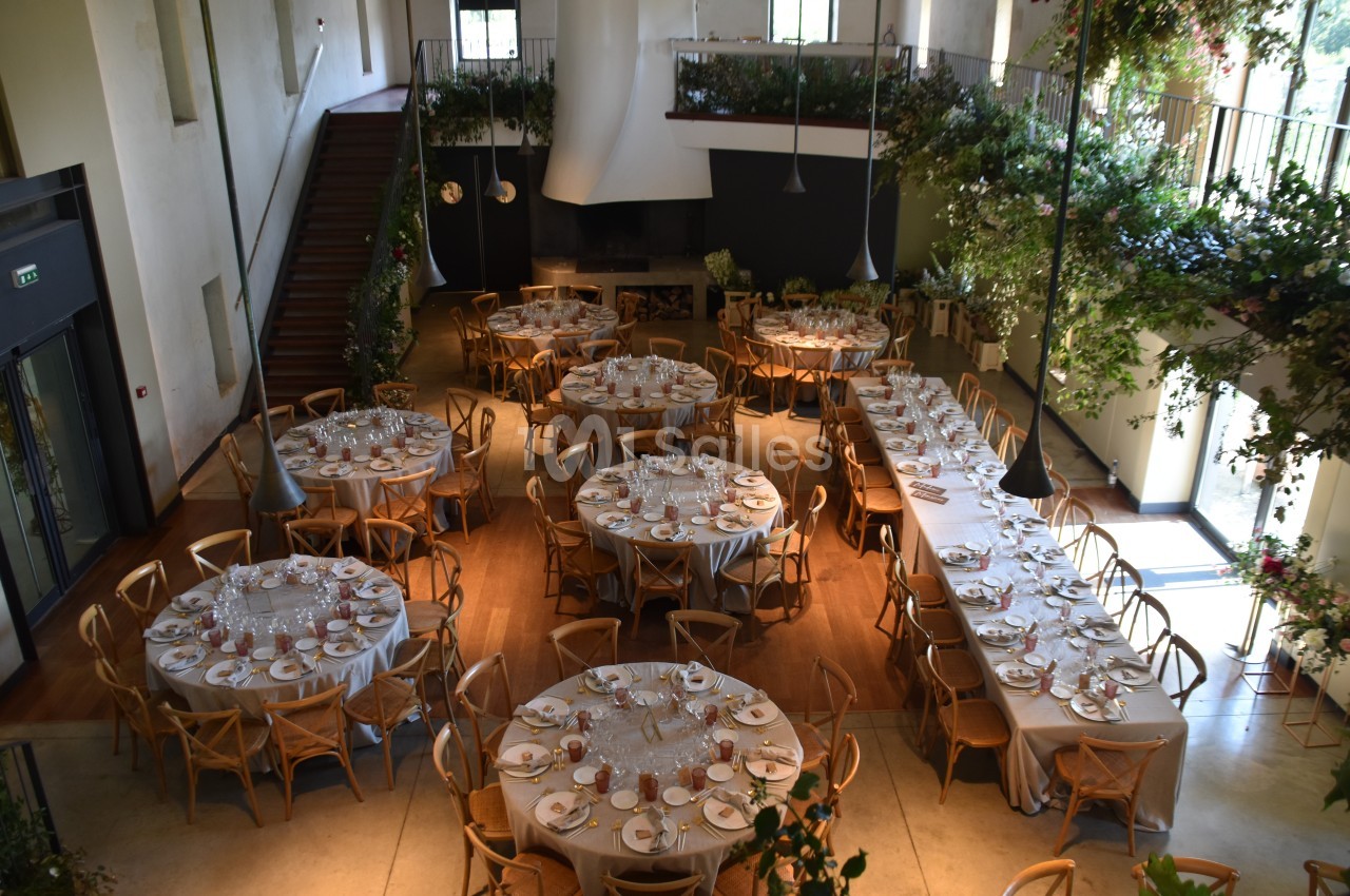 Salle de réception lumineuse avec tables rondes et rectangulaires dressées, entourées de chaises en bois.