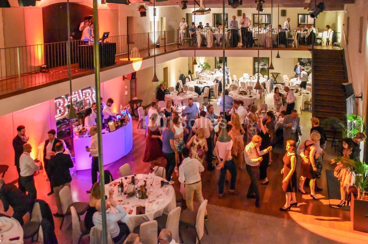 Salle de réception animée avec des invités dansant sur une piste, entourés de tables dressées et d'un bar éclairé.