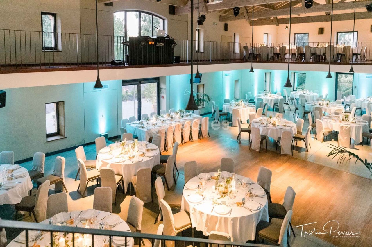 Salle de réception lumineuse avec tables rondes dressées pour un événement, décorée de fleurs et éclairée en bleu.
