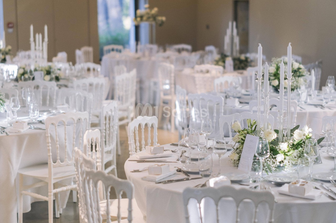 Salle de réception décorée avec des tables rondes, nappes blanches, chandeliers et arrangements floraux élégants.
