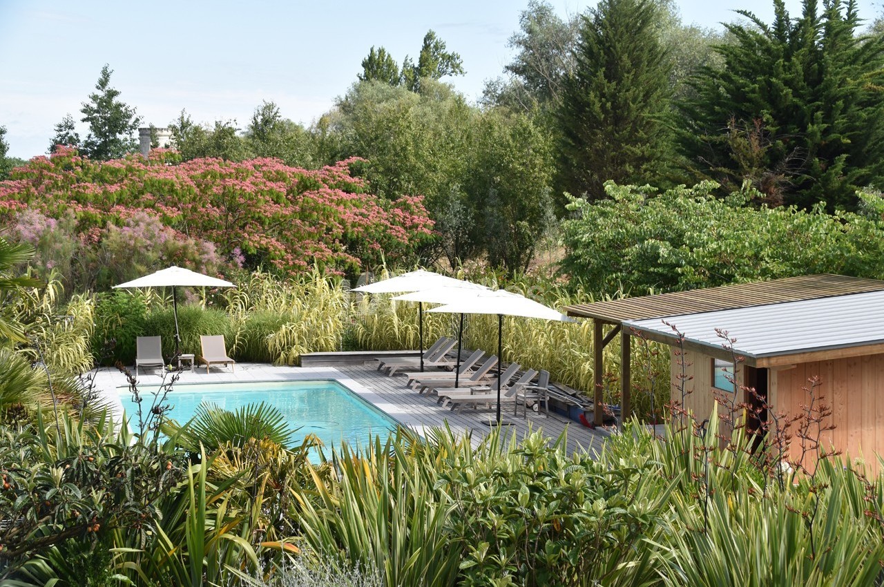 Piscine extérieure entourée de végétation luxuriante, avec chaises longues, parasols et un abri en bois.