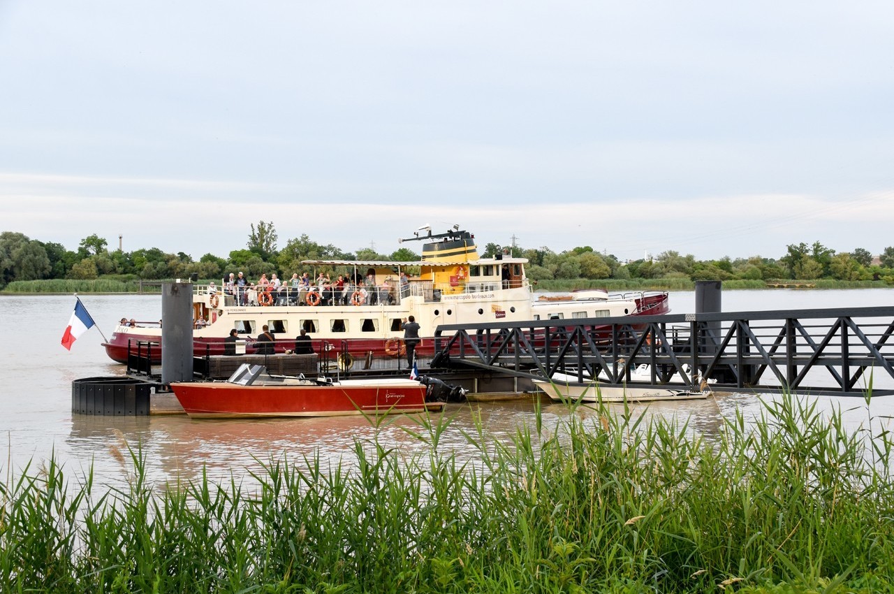 Bateau à passagers amarré à un ponton sur une rivière, avec un drapeau français et des arbres en arrière-plan.