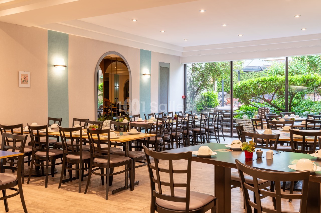 Salle lumineuse avec tables et chaises en bois, grandes baies vitrées donnant sur un jardin verdoyant.
