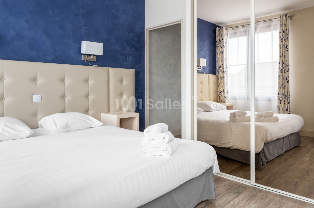 Chambre lumineuse avec un lit double, des draps blancs, des serviettes pliées et un grand miroir reflétant la fenêtre.