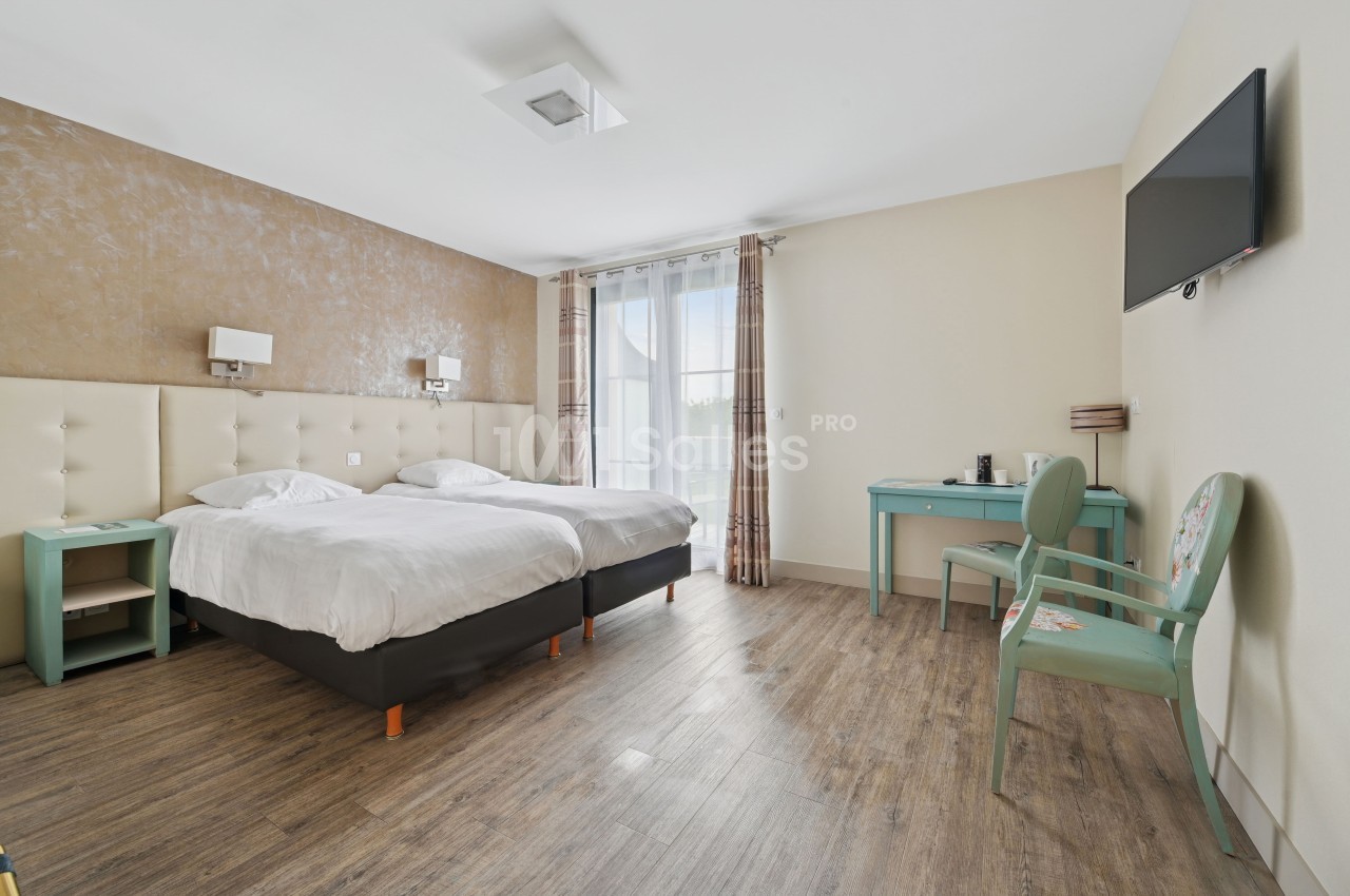 Chambre lumineuse avec deux lits simples, sol en bois, bureau, chaise, télévision murale et grande fenêtre avec rideaux.