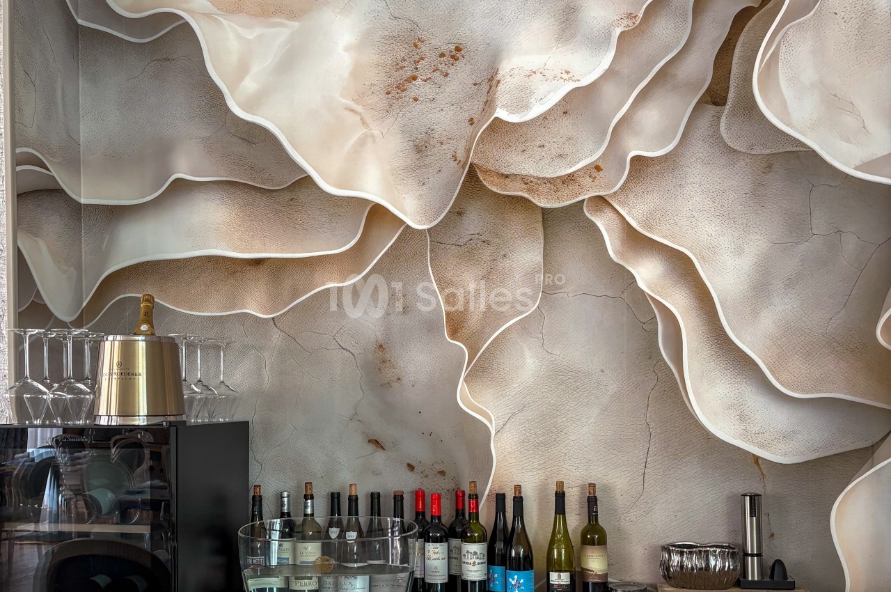 Mur texturé beige évoquant des formes organiques, avec un bar en premier plan contenant bouteilles et verres.