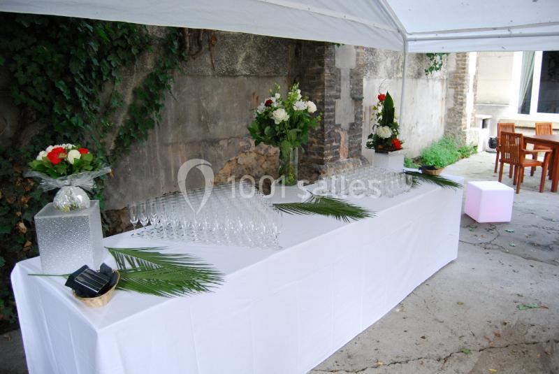 Table dressée sous une tente avec nappes blanches, verres alignés, bouquets de fleurs et feuilles décoratives.