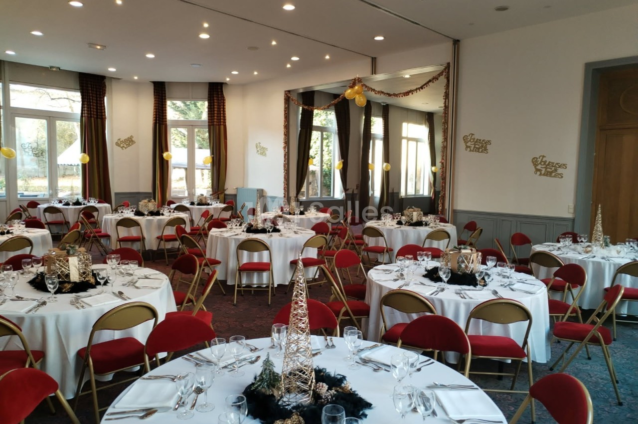Salle décorée pour un événement festif avec des tables rondes dressées, chaises rouges et ornements dorés.