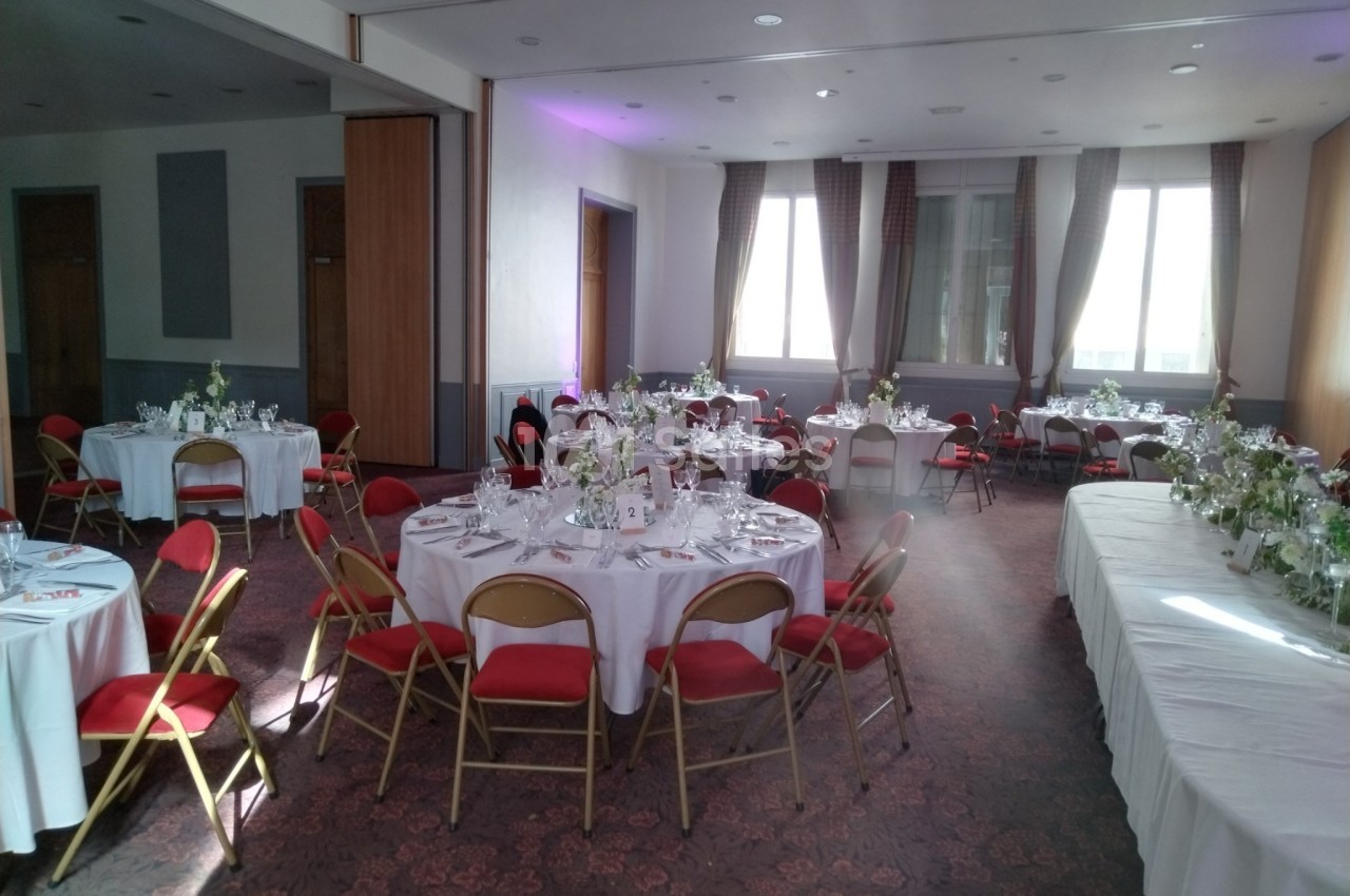 Salle de réception lumineuse avec tables rondes dressées, chaises rouges et nappes blanches, prête pour un événement.