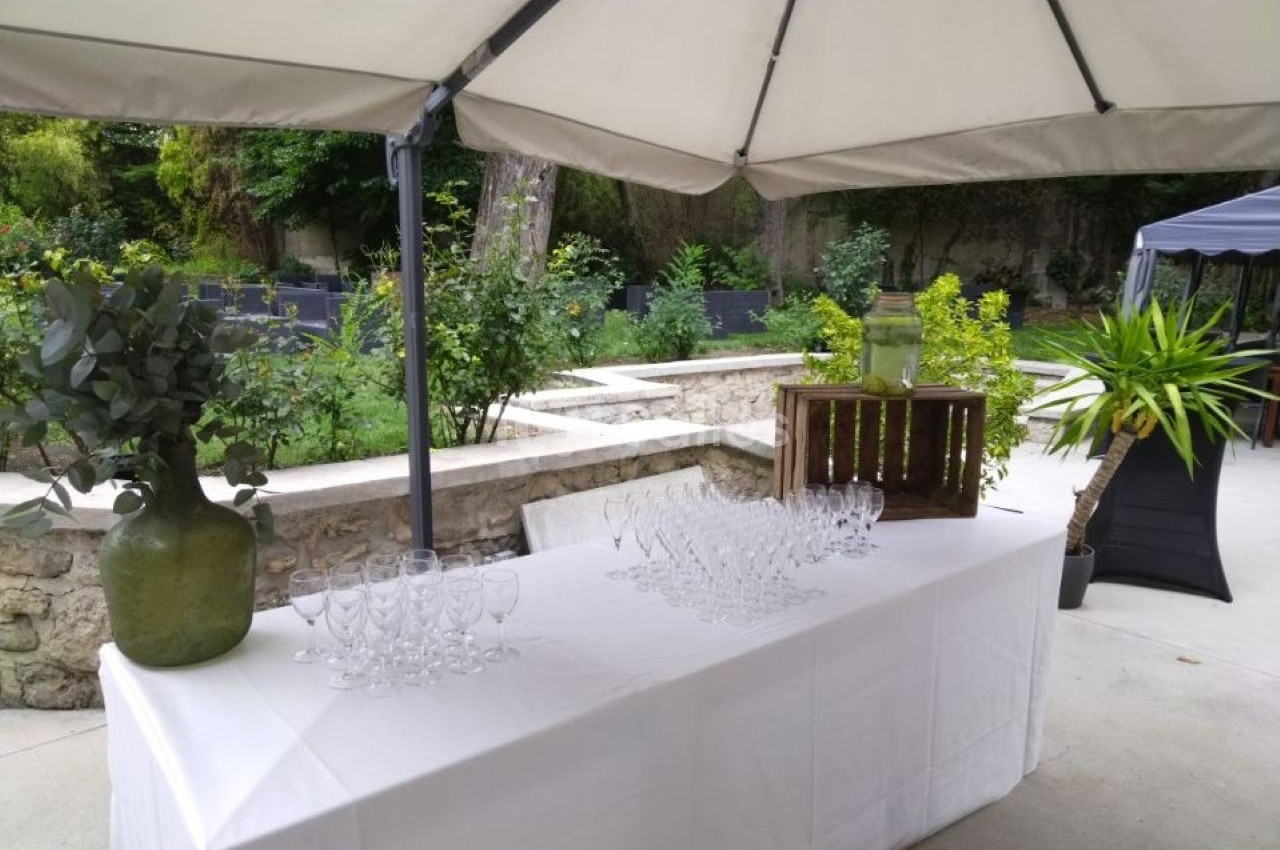 Table dressée en extérieur avec des verres, un vase vert et une caisse en bois sous un grand parasol.
