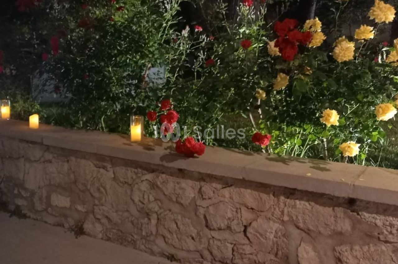 Des bougies allumées posées sur un muret en pierre devant des rosiers rouges et jaunes dans un jardin nocturne.