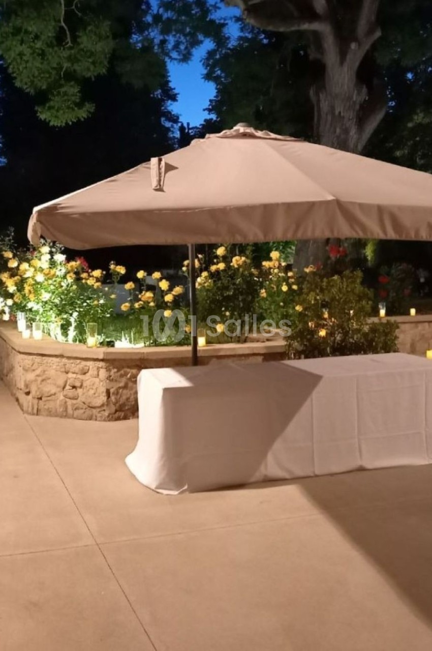 Parasol beige et table recouverte d'une nappe blanche dans un jardin éclairé en soirée, entouré de fleurs et de bougies.