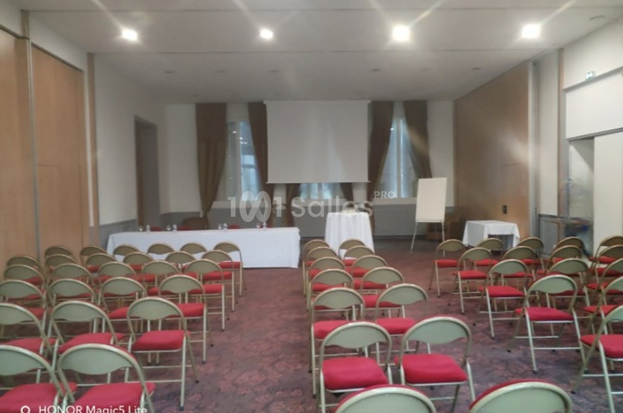 Salle de conférence avec des rangées de chaises rouges, une table avec micros et un écran de projection à l'avant.
