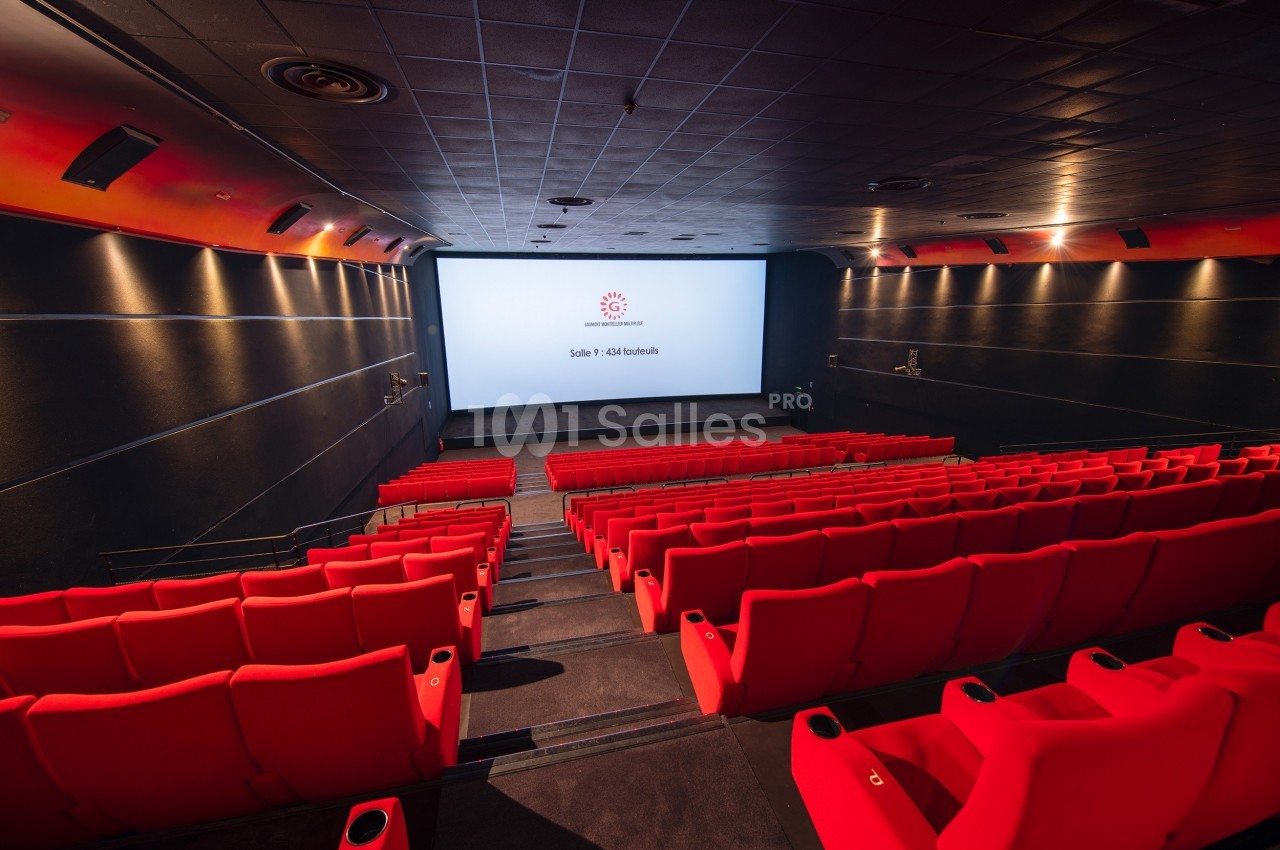 Salle de cinéma vide avec des rangées de sièges rouges et un écran blanc allumé au fond.