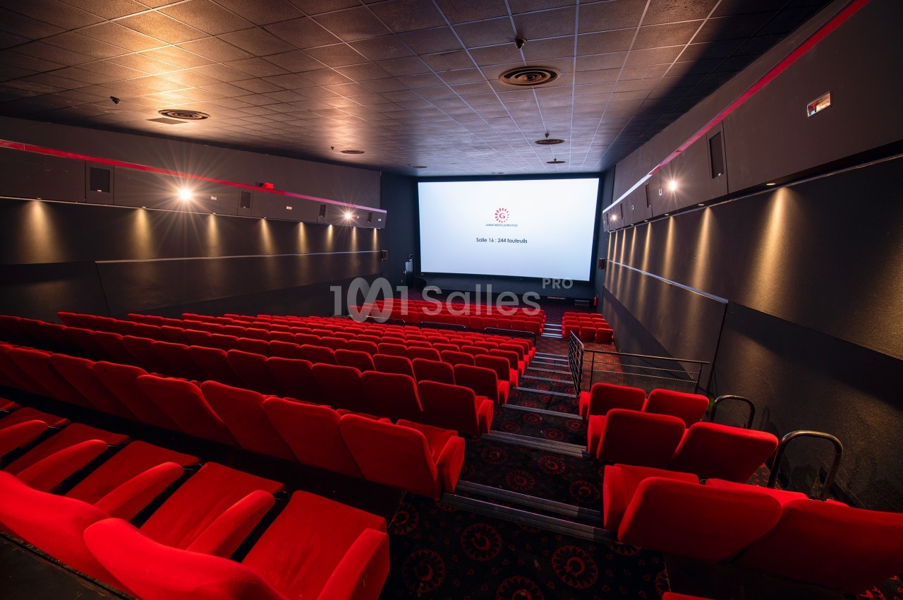 Salle de cinéma vide avec des rangées de sièges rouges et un écran blanc allumé au fond.