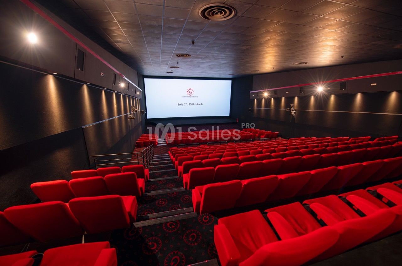 Salle de cinéma vide avec rangées de sièges rouges et écran blanc allumé dans un espace sombre.