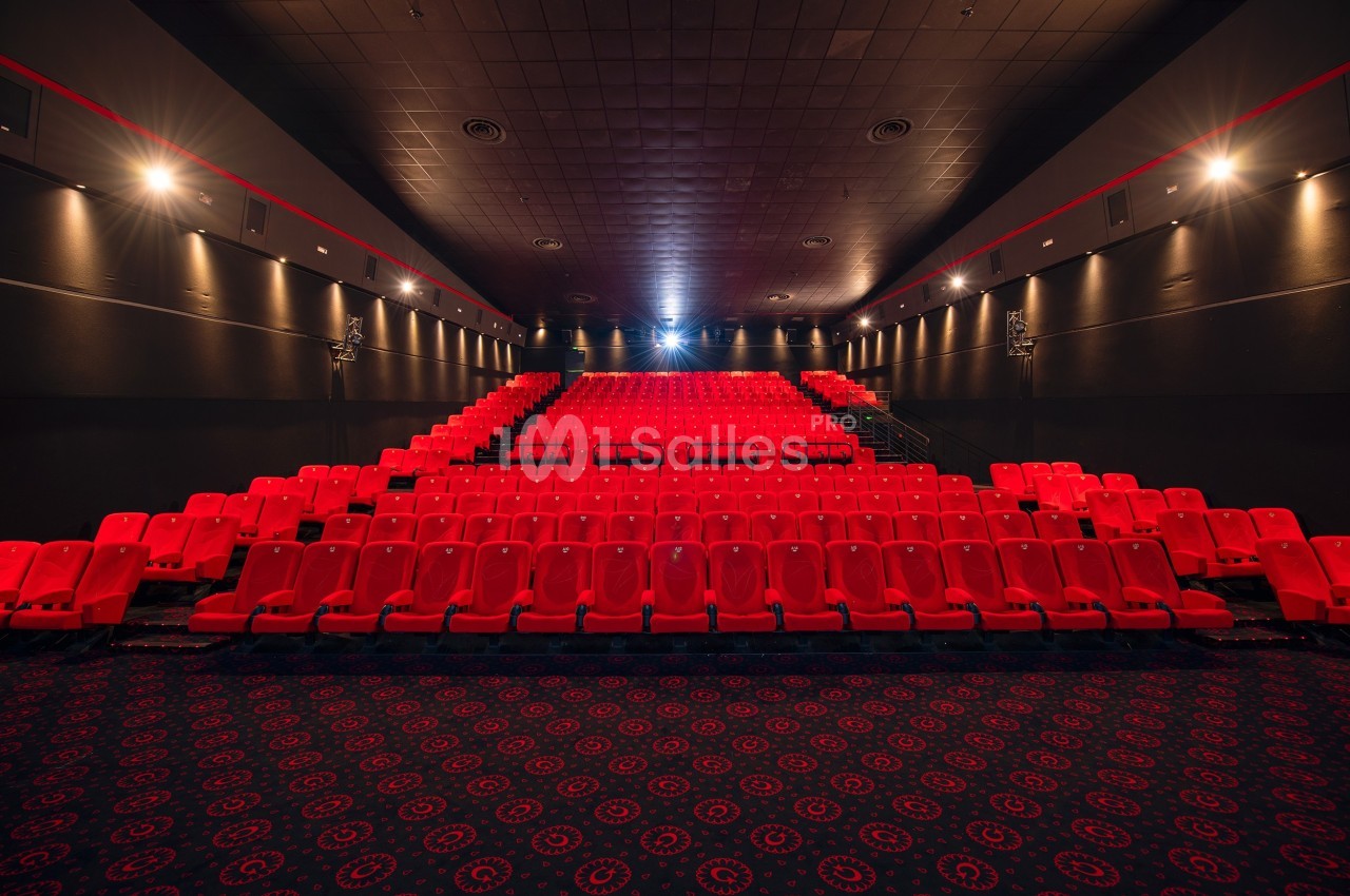 Salle de cinéma vide avec rangées de sièges rouges, éclairage tamisé et écran en arrière-plan.
