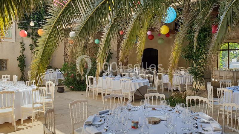 Salle de réception en plein air décorée de lanternes colorées, avec des tables rondes dressées sous des palmiers.