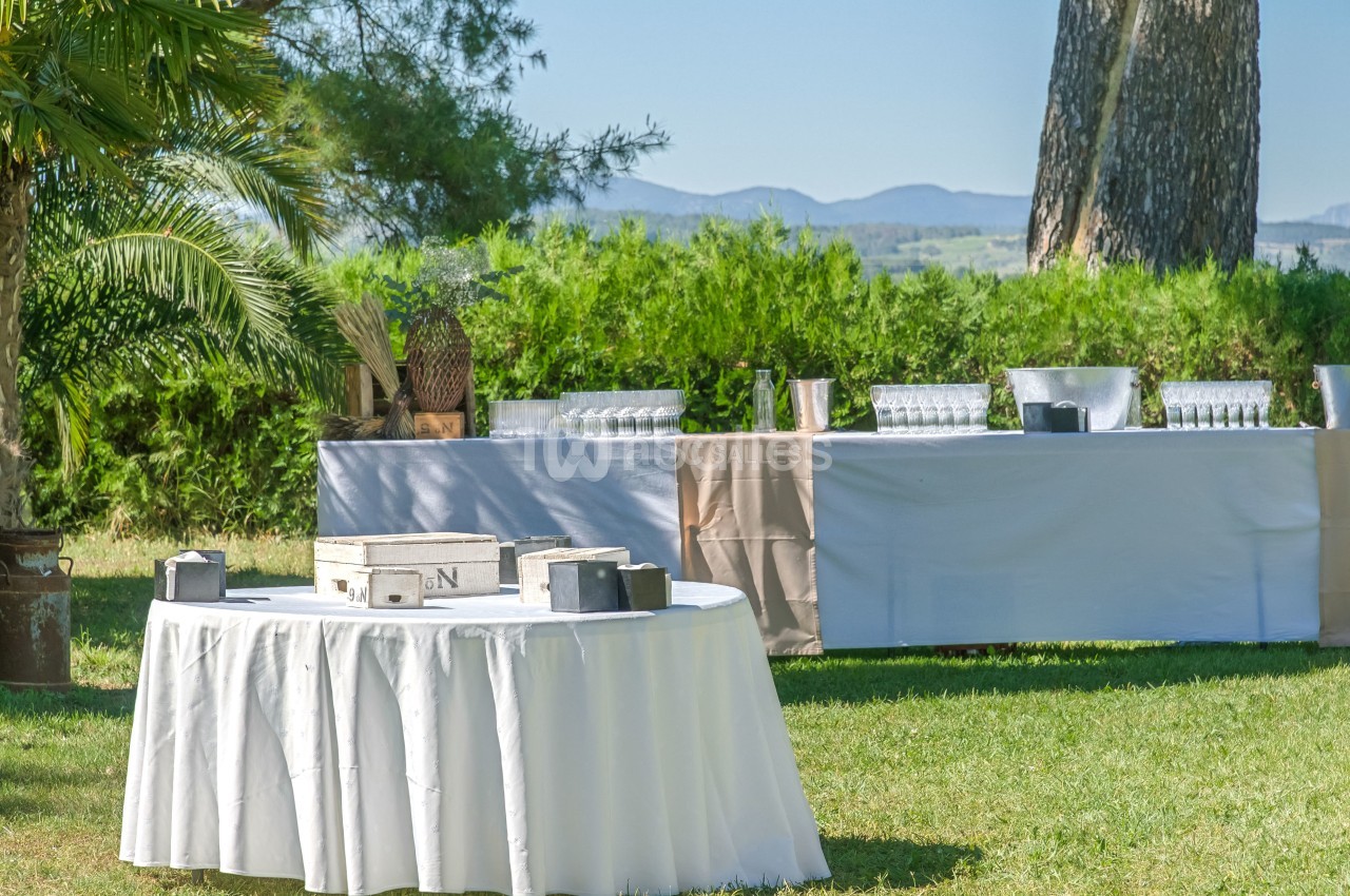 Tables dressées en extérieur avec nappes blanches, verres et décorations, entourées de verdure et d'arbres.