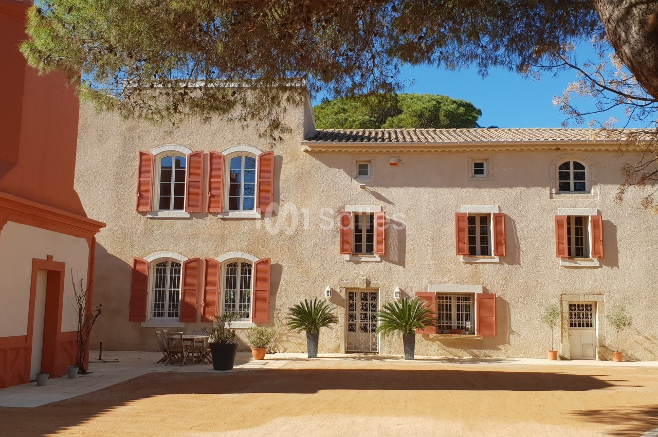 Façade d'une maison provençale avec volets rouges, arbres et terrasse ensoleillée sur une cour en gravier.