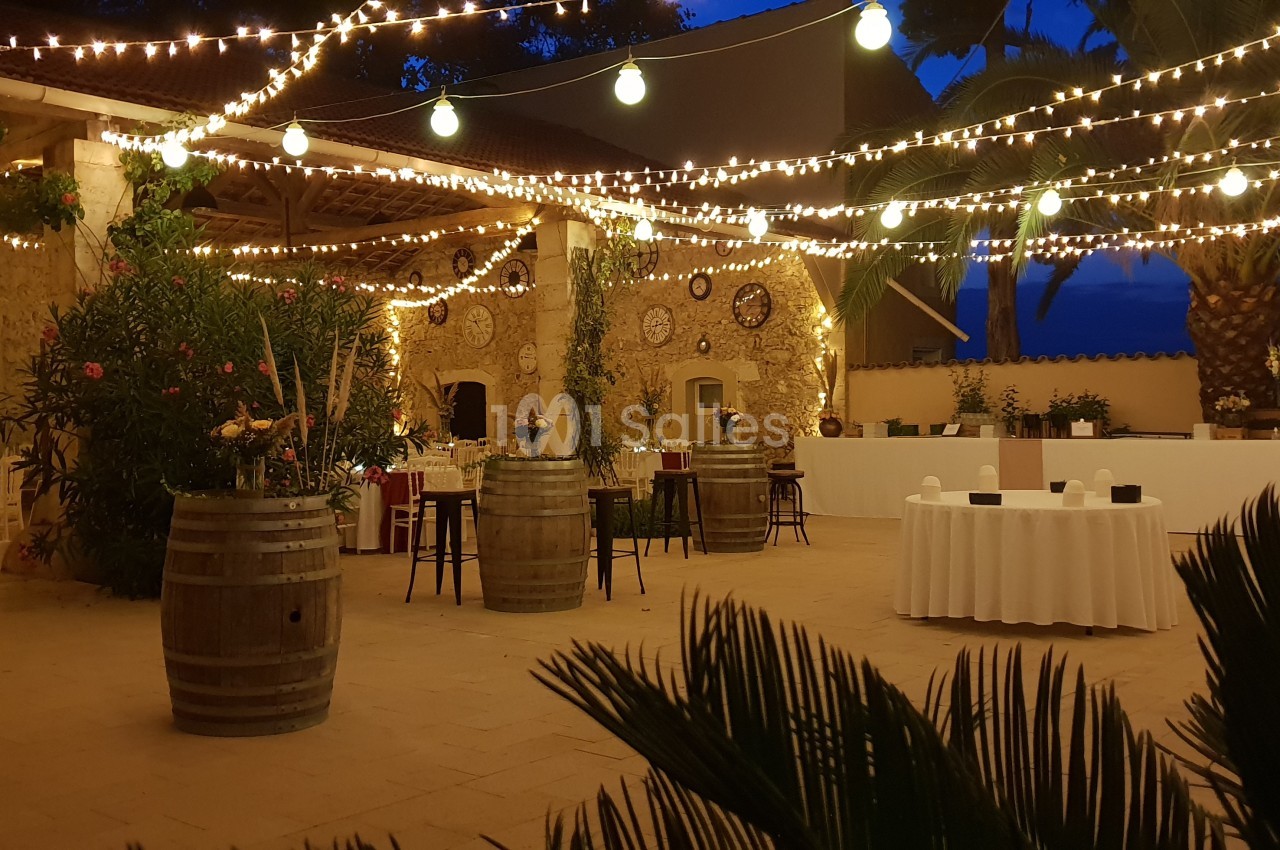 Cour extérieure décorée de guirlandes lumineuses, tables dressées et tonneaux, ambiance festive en soirée.