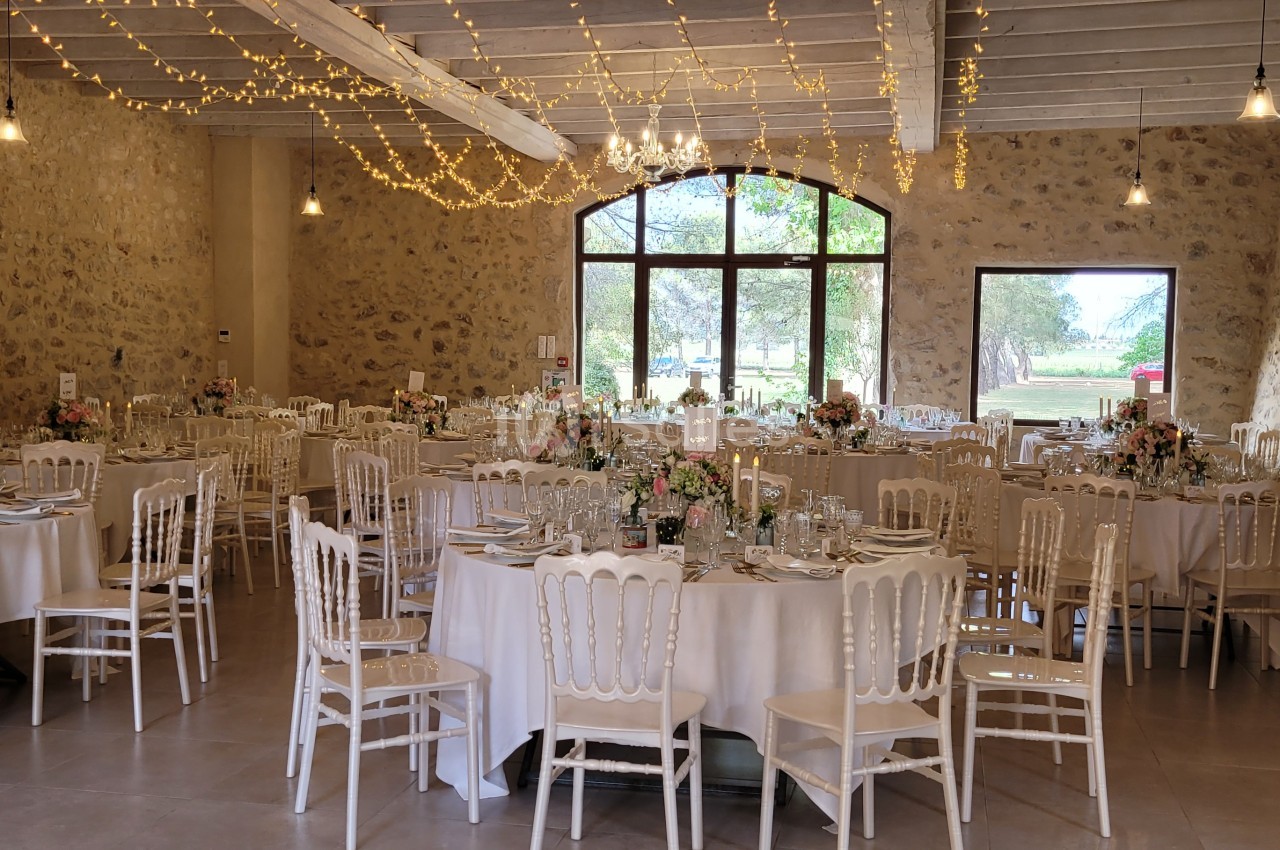 Salle de réception décorée avec des tables rondes, des chaises blanches et des guirlandes lumineuses suspendues.