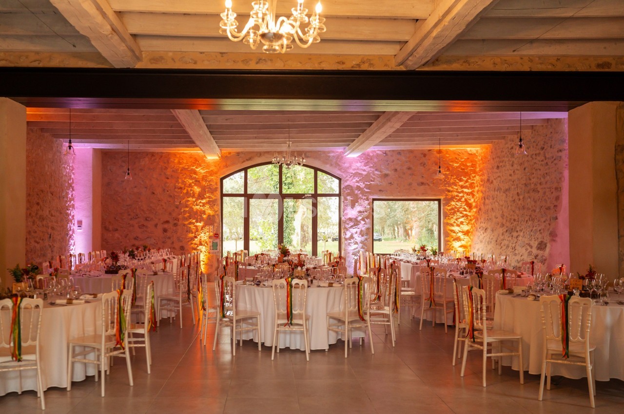 Salle de réception décorée avec des tables rondes dressées, éclairage chaleureux et grandes baies vitrées donnant sur un…