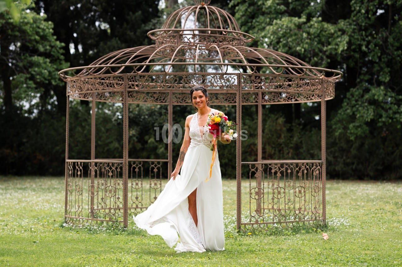 Femme en robe blanche tenant un bouquet coloré devant une structure métallique décorative dans un jardin verdoyant.