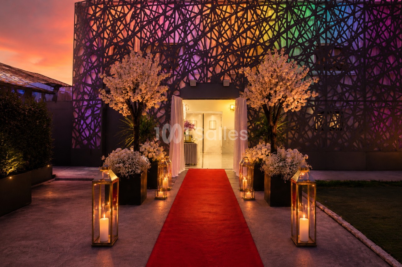 Entrée décorée avec un tapis rouge, des lanternes allumées et des fleurs, devant un bâtiment illuminé de lumières colorées.