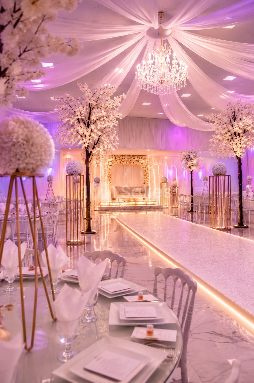 Salle de réception élégante décorée de fleurs blanches, éclairage rose et lustre central sous un plafond drapé.