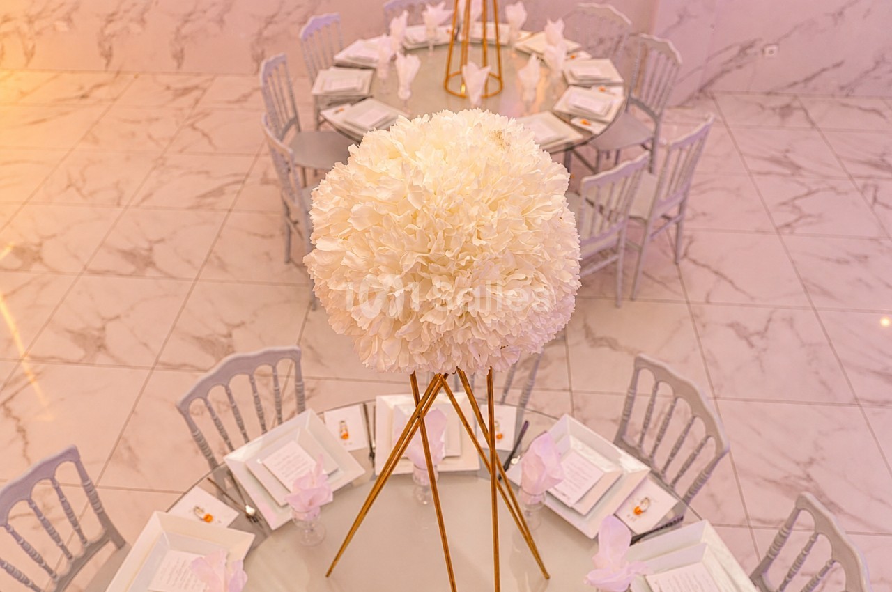 Centre de table floral blanc sur trépied doré, entouré de tables rondes dressées avec nappes blanches et chaises grises.