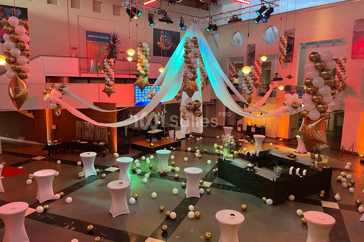 Salle décorée pour un événement avec ballons dorés et blancs, tables hautes et éclairages colorés.