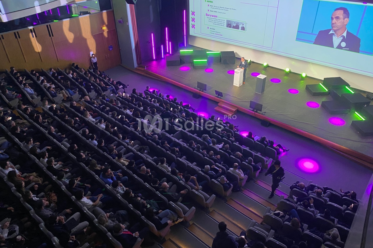 Vue d'une conférence dans un auditorium, avec un orateur sur scène et un public nombreux assis.